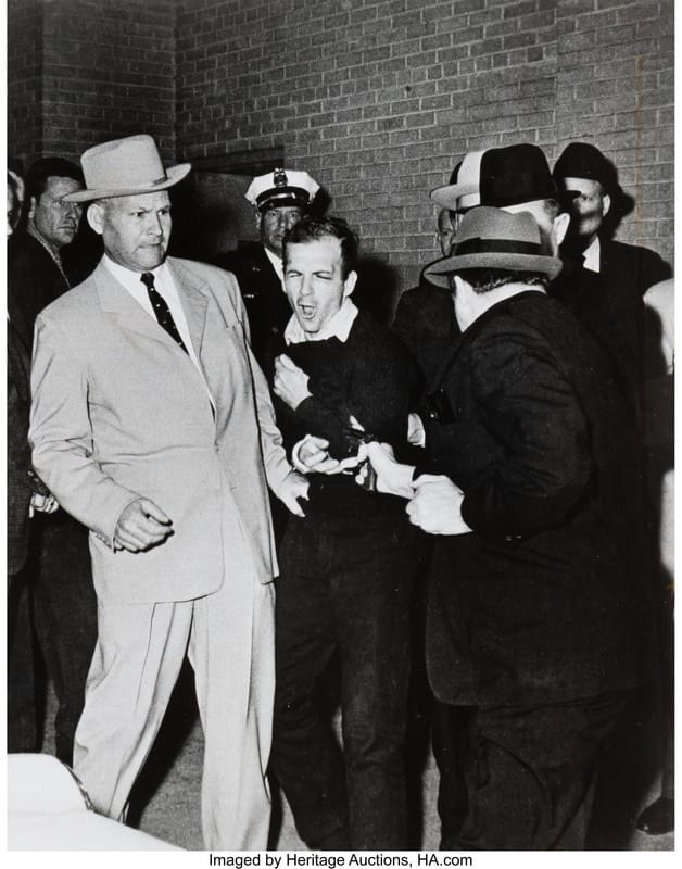 Robert Hill Jackson - 1EarthMedia Black-and-white photo of a man in a dark sweater being restrained by two men in suits and hats as others look on near a brick wall, tense scene.