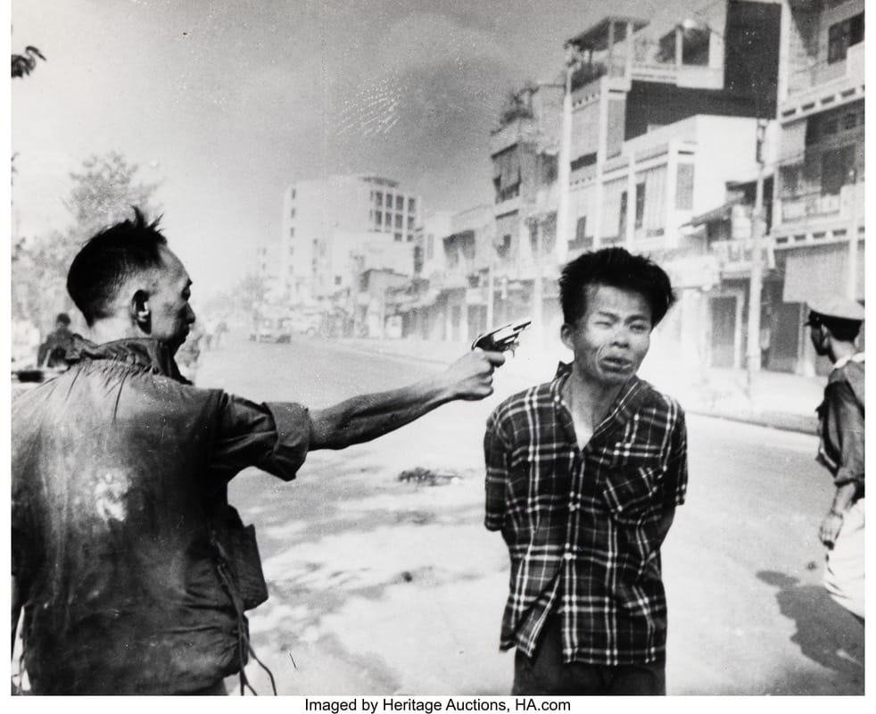 Eddie Adams - 1EarthMedia Man in a plaid shirt stands with hands behind his back as another man points a gun at him on a smoky, damaged street.