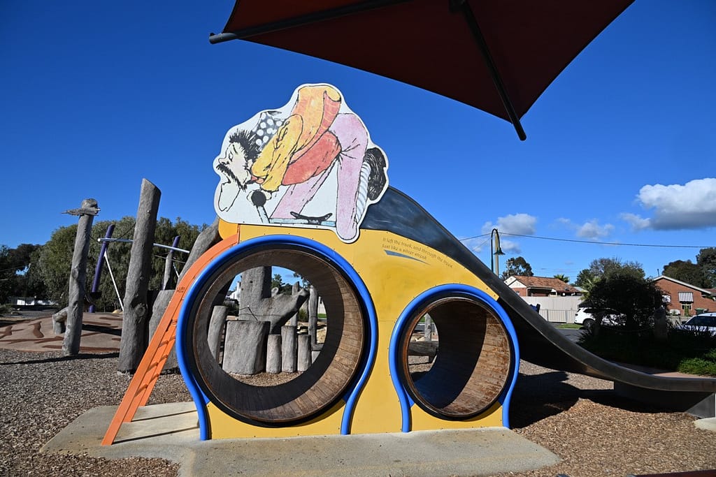 MulgaBill--6a - 1EarthMedia Colorful playground structure with two circular blue-trimmed tunnels and a curved slide under a clear blue sky.