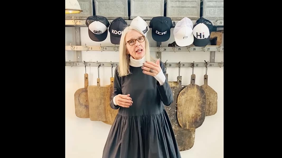 Diane Keaton’s Home: The Beauty of a Life Collected Woman with blonde hair and glasses speaks, wearing a dark dress in a workshop with hanging cutting boards and hats behind her