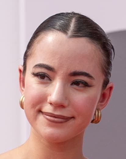 Close-up portrait of a woman with dark hair slicked back, wearing gold hoop earrings and subtle makeup, smiling softly.