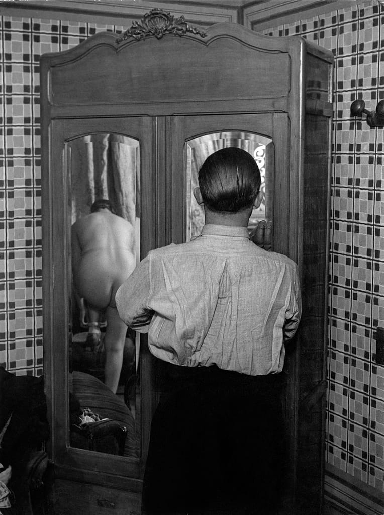 La toilette dans un hôtel de passe rue Quincampoix 1 c - 1EarthMedia Man in a shirt stands with his back to the camera in front of a vintage wooden armoire; a nude person is visible in the mirrored door reflection.