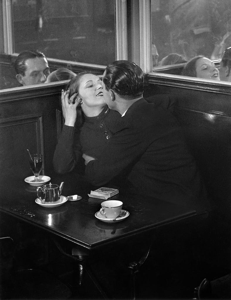 Brassaï Couple d_amoureux dans un petit café parisien Place d_Italie Paris c - 1EarthMedia Vintage couple kissing in a dark cafe booth, with a glass of wine and teapot on the table, cigarette pack nearby and mirrors reflecting their embrace.