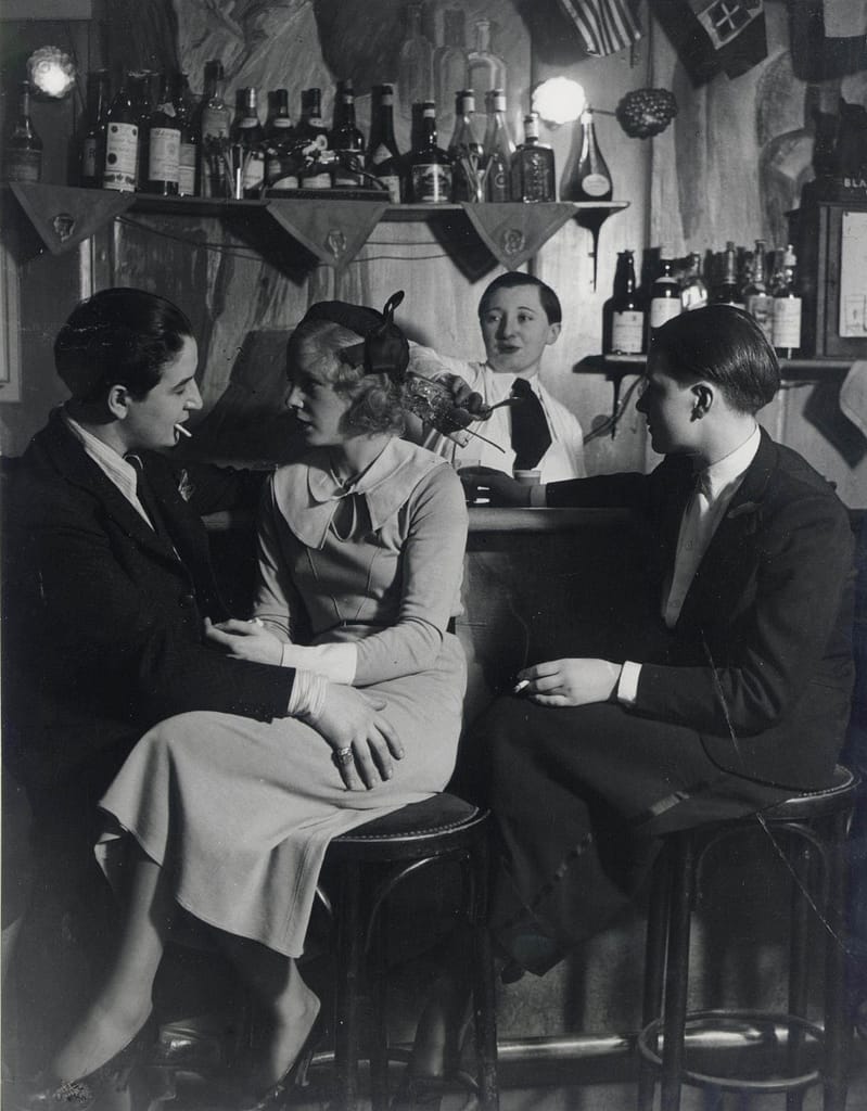Brassai-auMonocle003 - 1EarthMedia Four patrons sit at a vintage bar: a man and woman on the left share a moment while a second man sits at right; bartender pours a drink behind them, bottles lining the shelf.
