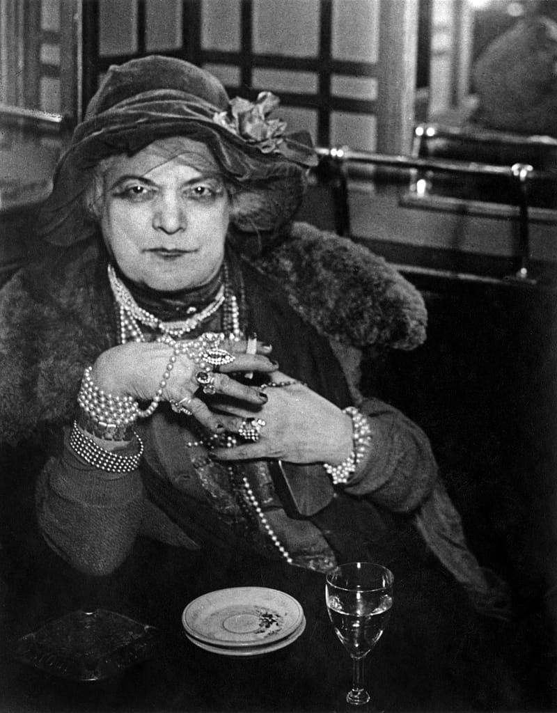 Brassaï La Môme Bijou au Bar de la Lune Montmartre c - 1EarthMedia Elegant woman in a vintage hat and fur stole, adorned with pearls, sits at a dining table with hands clasped; plate and wine glass visible.