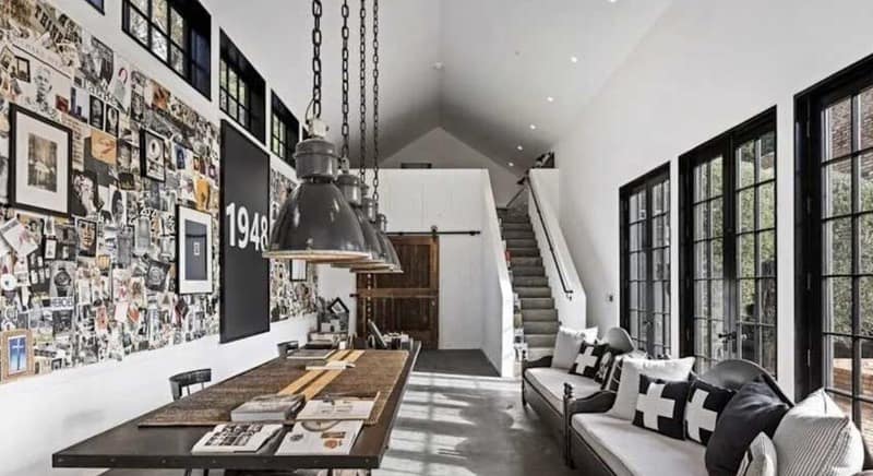 Diane Keaton The Wall - 1EarthMedia Bright industrial loft with a collage wall, long wooden table, and a gray sofa area under large pendant lights by tall windows.