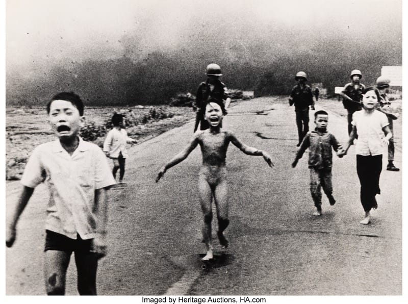 Nick Ut - 1EarthMedia Children run toward the camera along a road, with soldiers and smoke visible in the background (black-and-white photo).