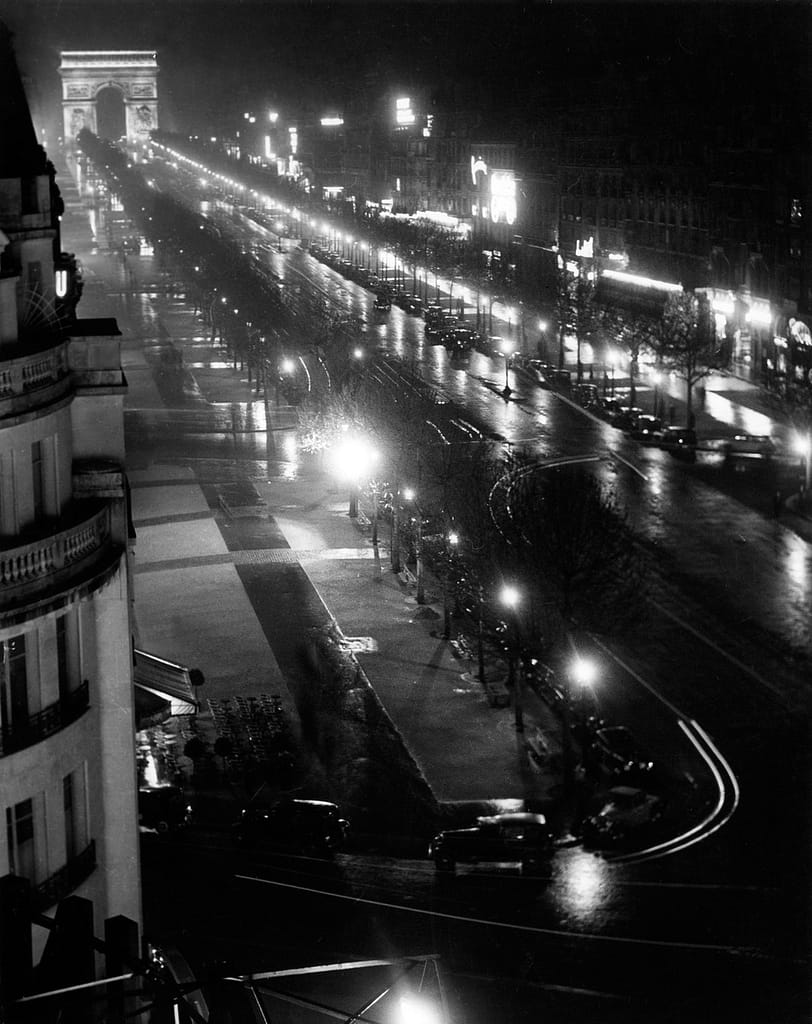 Arc de Triomphe et Champs Elysées ca - 1EarthMedia Night view of a wet Paris street lined with trees and bright signs, with the Arc de Triomphe visible in the distance.