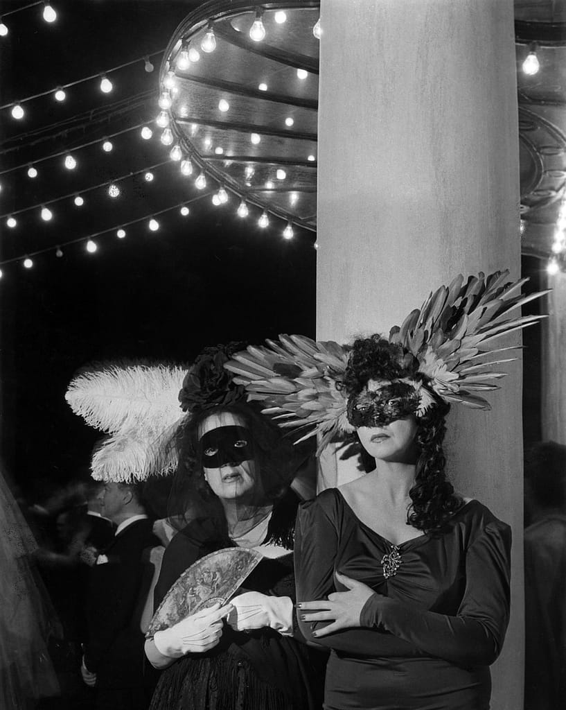 Le bal des têtes au Pré Catelan -ML - 1EarthMedia Two women in elegant gowns wearing feathered masquerade masks at a gala under string lights, posing together by a pillar at night option 1