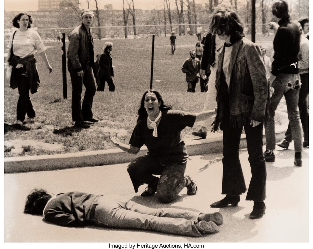 John Filo - 1EarthMedia A woman kneels beside a person lying face down on the pavement while others stand nearby in a park, appearing distressed or surprised.